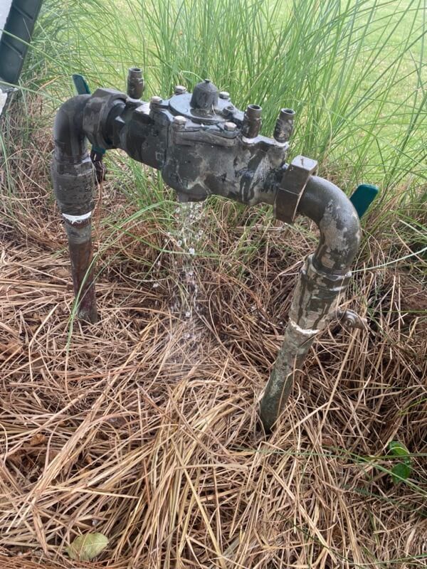Leaking outdoor metal pipe system over dry grass and straw, surrounded by green foliage.