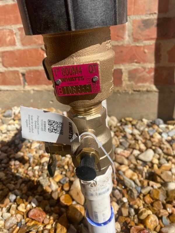 Brass valve with label "WATTS 800M4 QT," outdoors by brick wall and gravel.