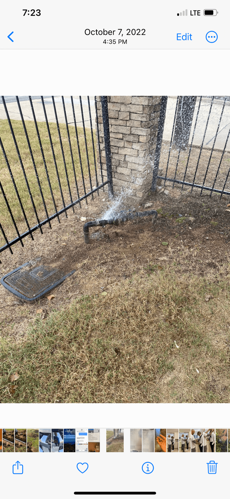 Sprinkler pipe leaking water near metal fence and brick pillar on dry grass and exposed soil.