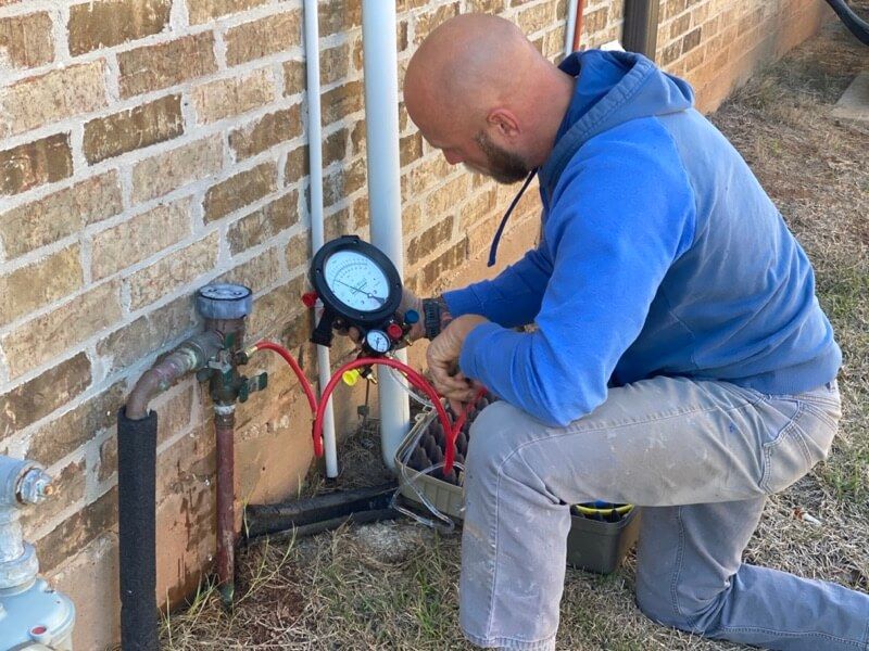 Man kneeling by brick wall inspecting pressure gauge with red hoses, suggesting repair work.