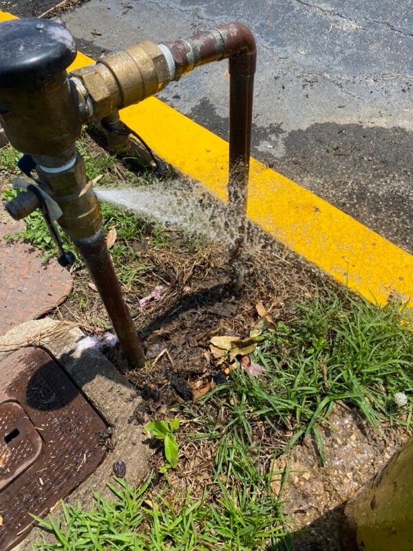 Leaking water pipe with valve spraying water on wet ground near yellow-lined pavement.