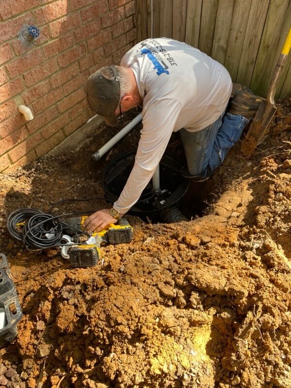 Person kneels in excavation near brick wall, working on pipes with tools and cables nearby.
