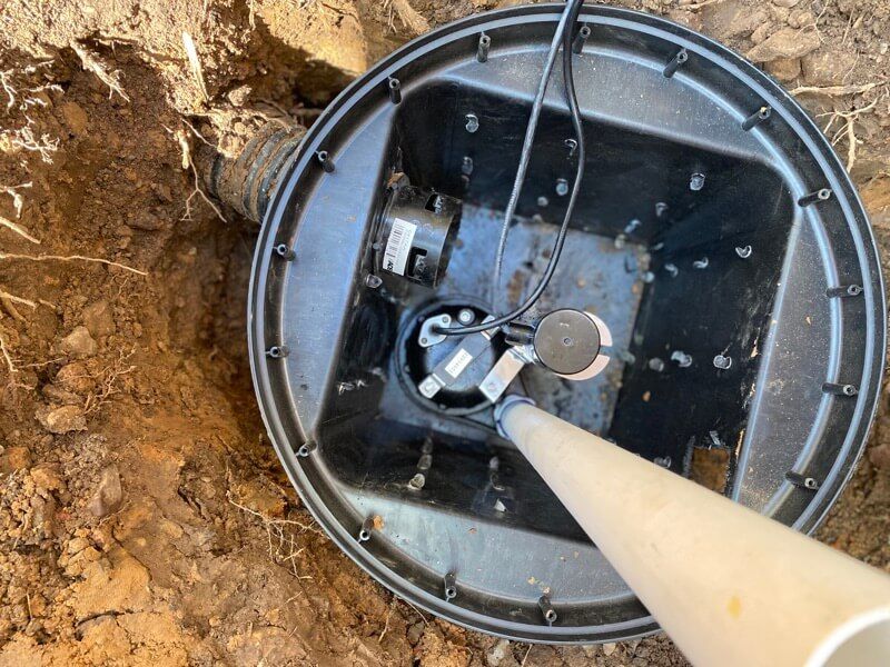 Black underground junction box with white conduit pipe and cables, surrounded by soil.