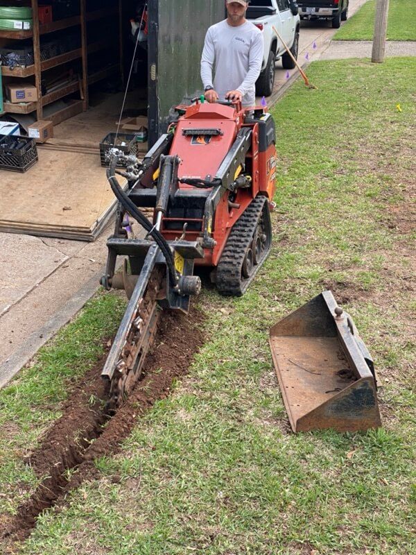 Person using a tracked trencher on grass with truck and trailer in background.