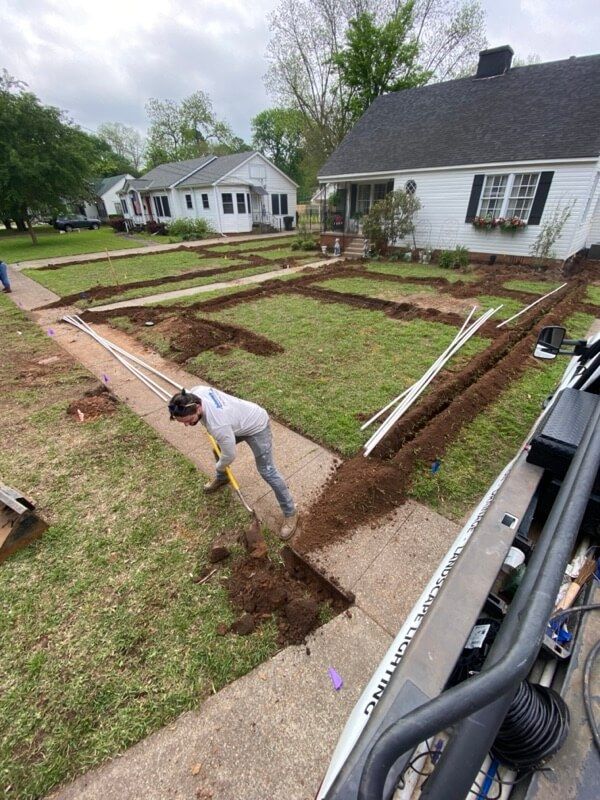 Person digging trench in suburban front yard with pipes and outlined trenches visible.