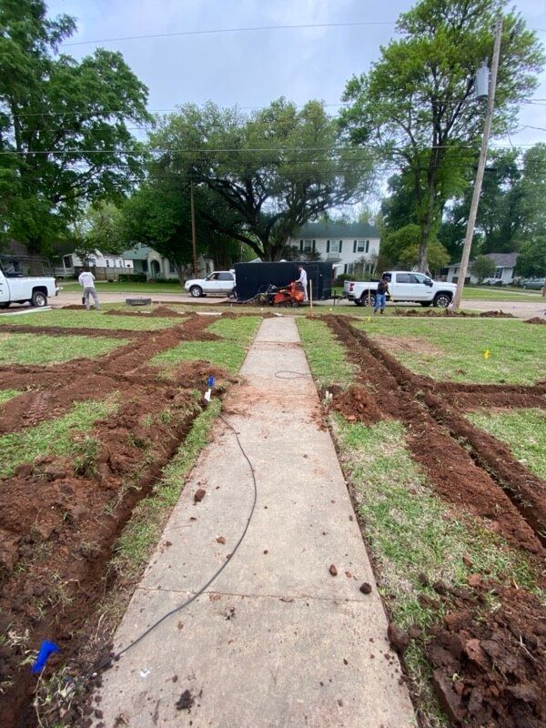 Pathway with trenches in grassy area, trees and parked trucks, workers present.