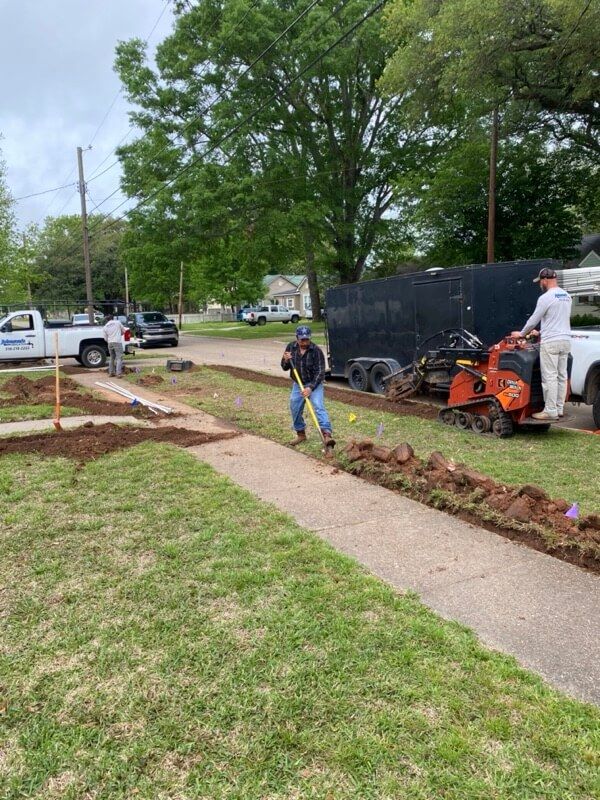 Man digging trench along sidewalk; mini track loader and trailer nearby.