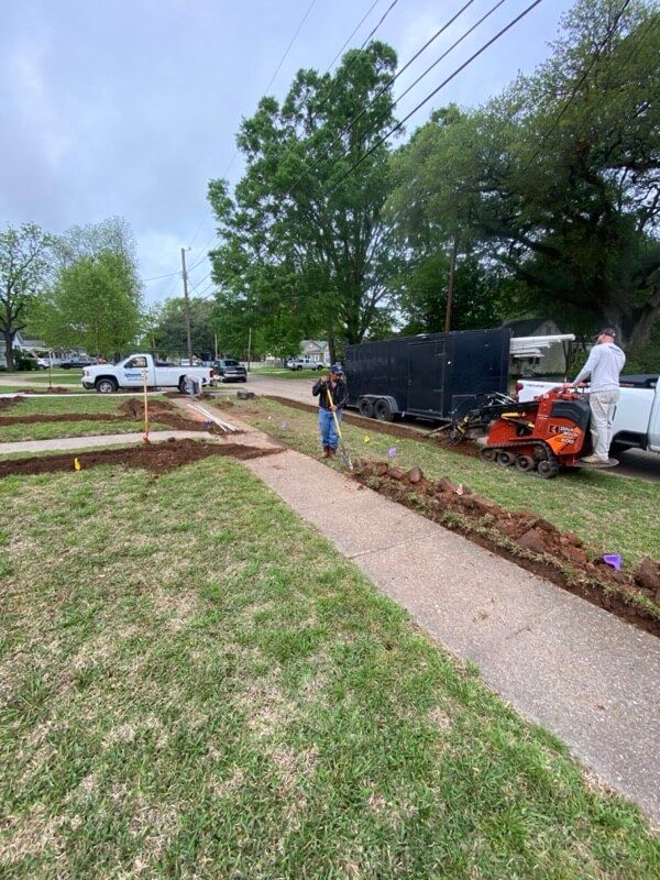 Person landscaping next to a sidewalk, with excavator, truck, and trailer nearby; trees visible.