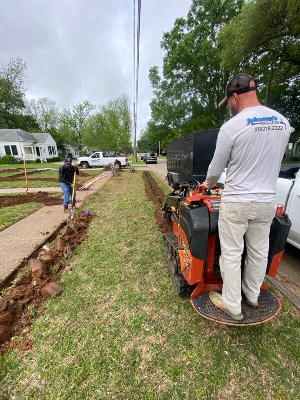 Workers landscaping on residential street with machinery, shovel, trucks, and trees visible.