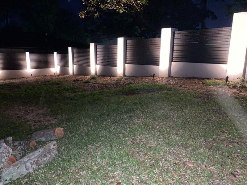 Yard at night with lit fence panels, grassy ground, logs, and trees in the background.