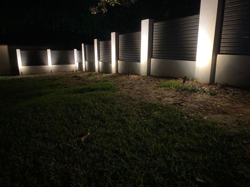 Modern fence illuminated by ground lights with grass and exposed soil in the foreground.