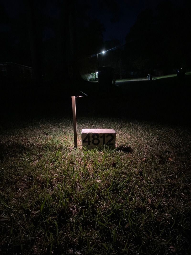 Number plaque "4812" lit by fixture on grass at night; dim suburb street with lamp in background.