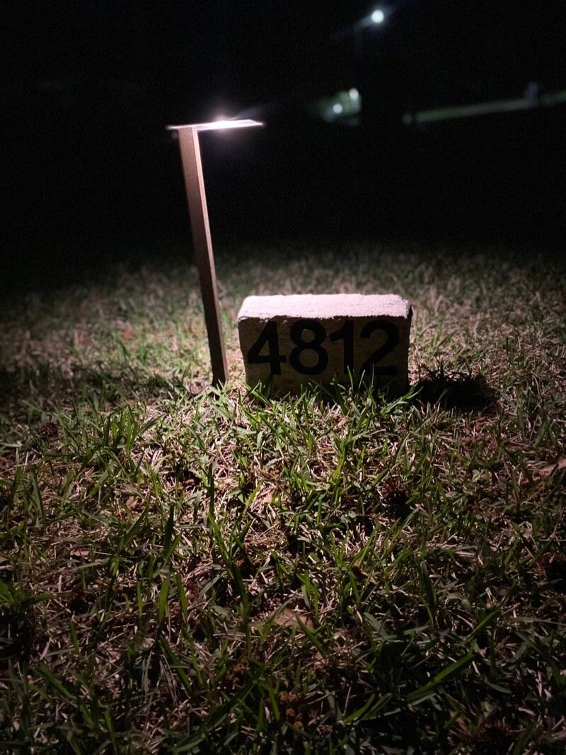 Wooden post with light illuminates grass, brick with number 4812, dark background.