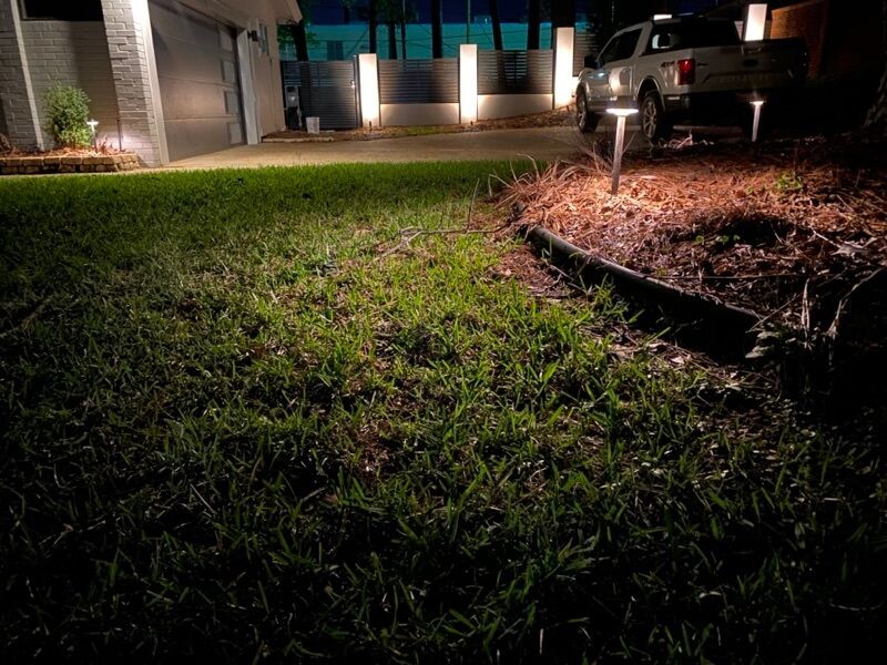 Nighttime suburban driveway scene, lit with pathway lights; vehicle parked near a garage.